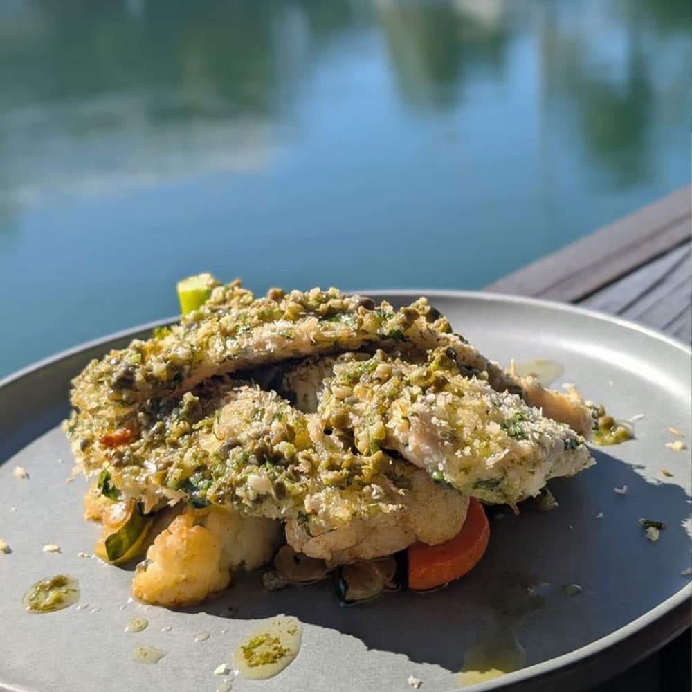 A plated fish dish topped with herbs and served with vegetables by a lakeside.