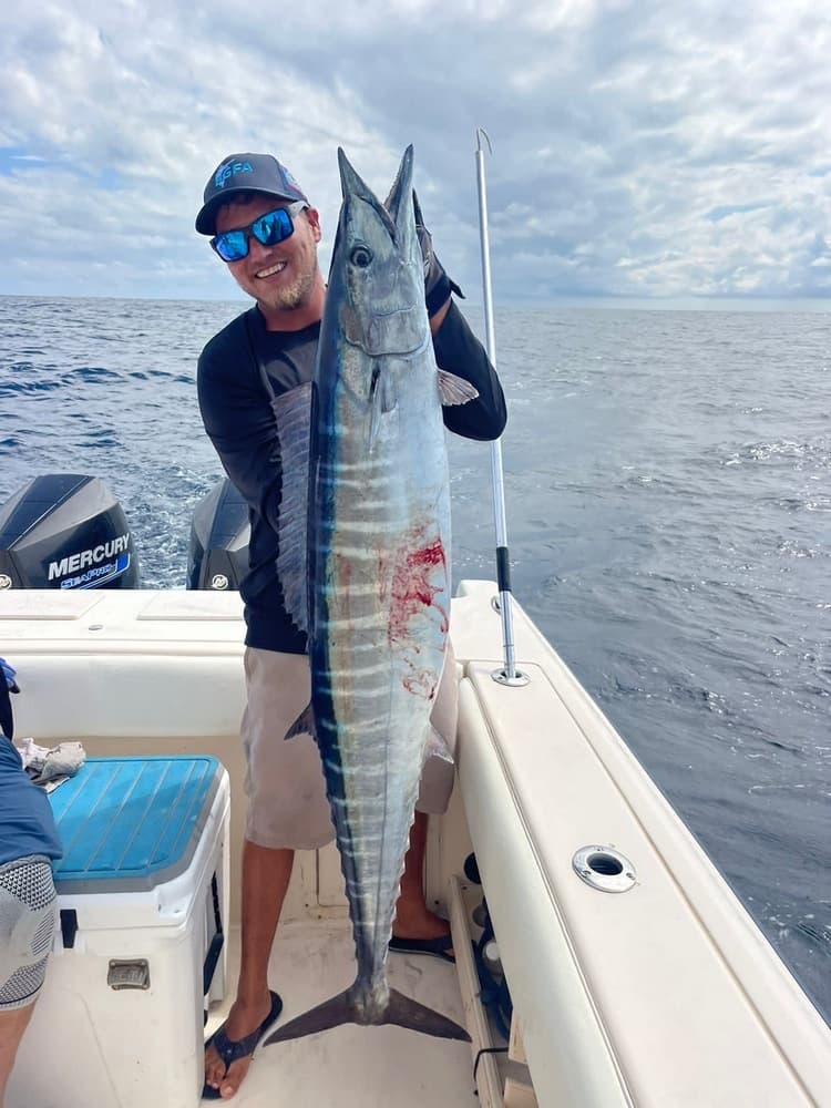 A smiling fisherman holds a large fish on a boat with ocean and cloudy sky in the background.