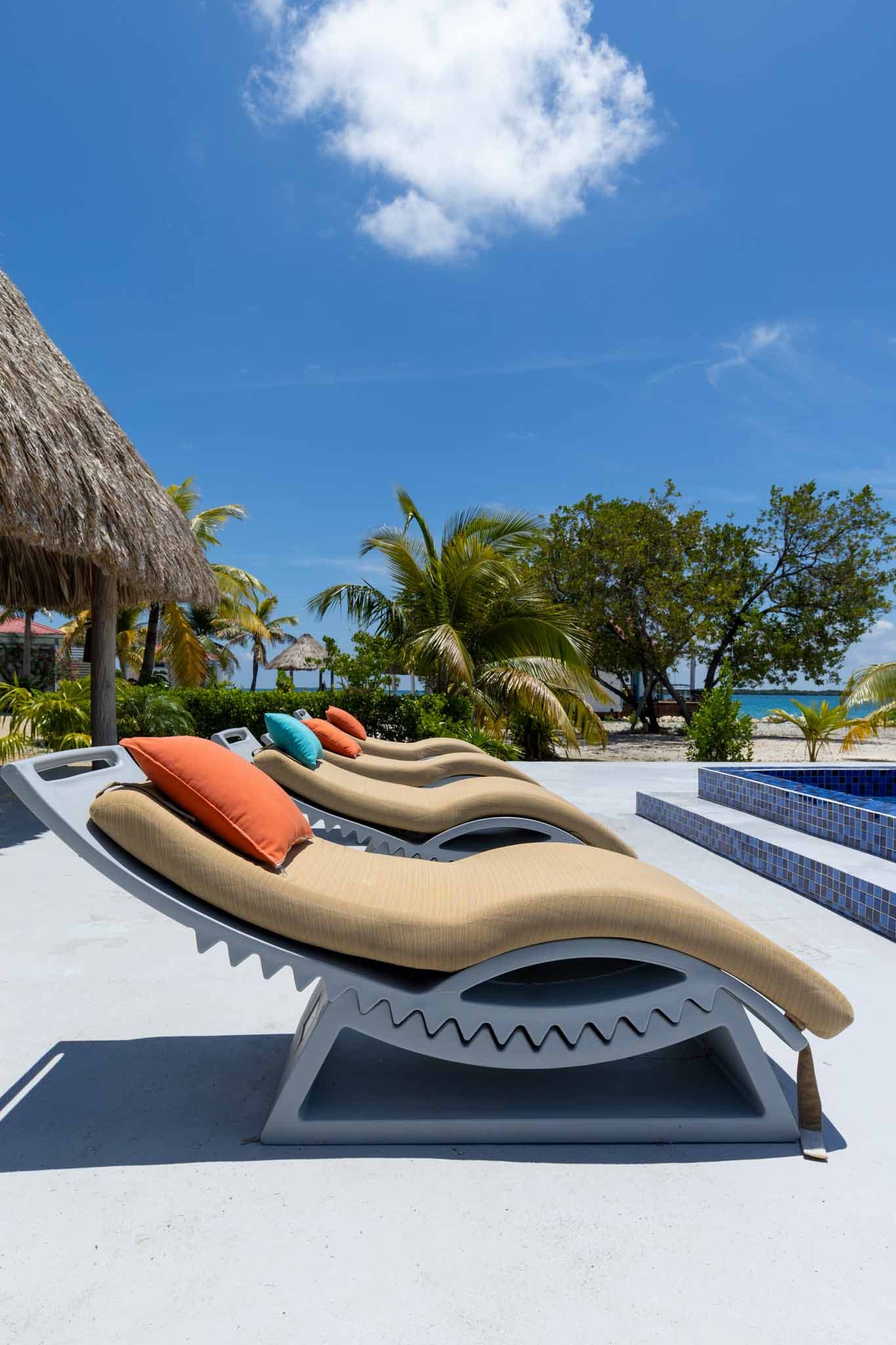 Three lounge chairs with colorful cushions are positioned by a poolside surrounded by palm trees and a clear blue sky.