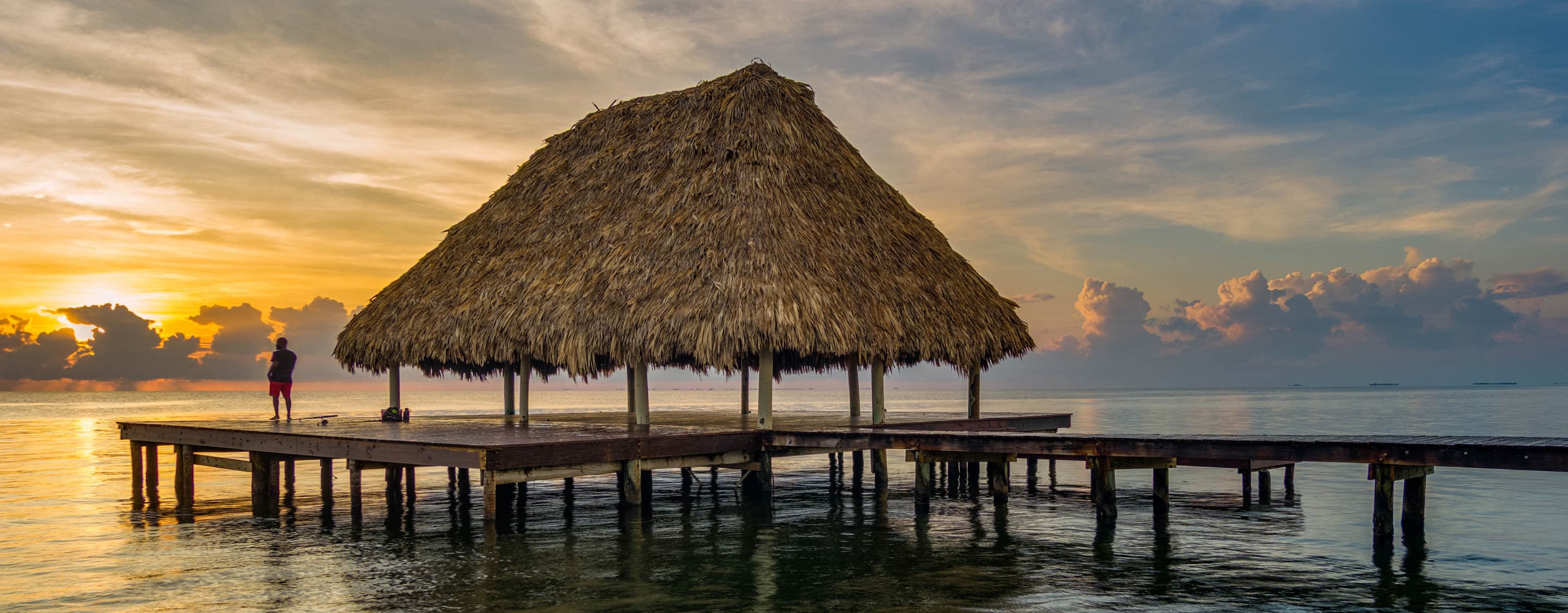A thatched-roof gazebo on a pier overlooking calm waters during sunset.
