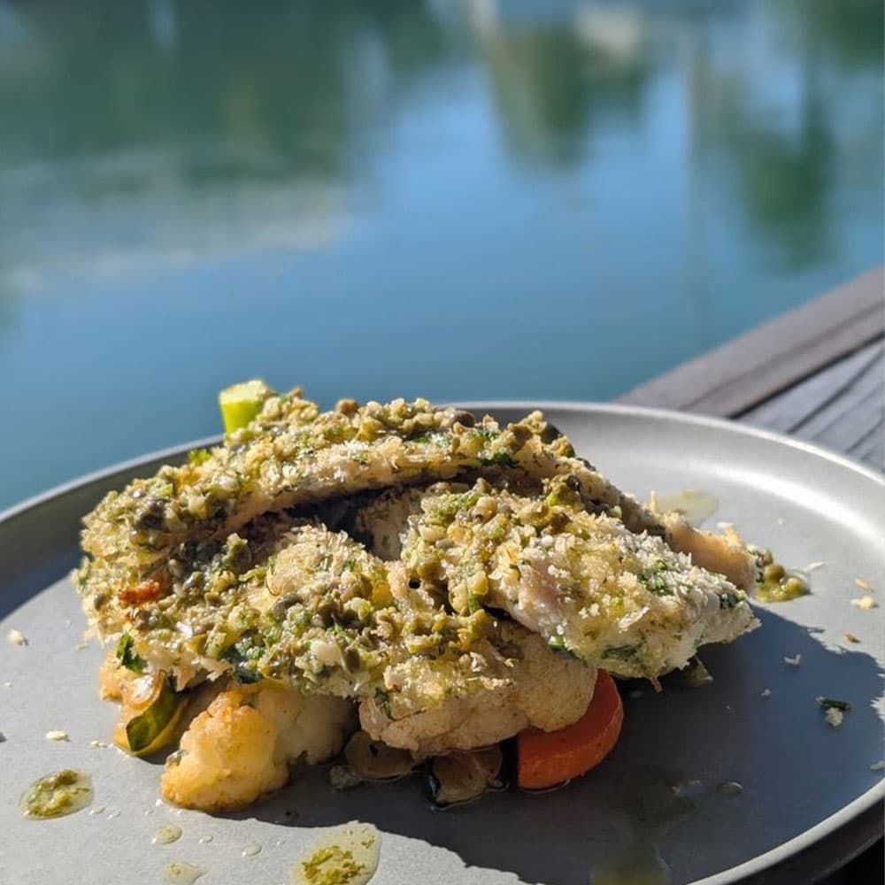 A plate of fish topped with a green herb mixture, served on a bed of vegetables, with a serene water backdrop.