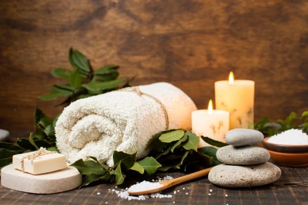 A rolled towel, candles, stones, and soap are arranged on a wooden surface with green leaves and salt.