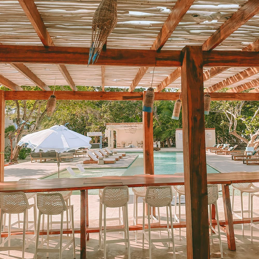 A view of a sunny pool area with loungers from an open bar under a wooden pergola.