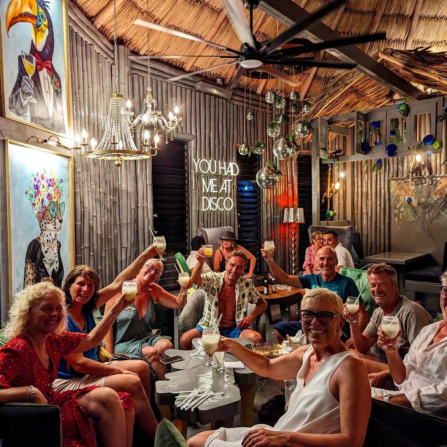 A group of people cheerfully raising their drinks in a lively, dimly lit bar adorned with vibrant artwork and decorations.