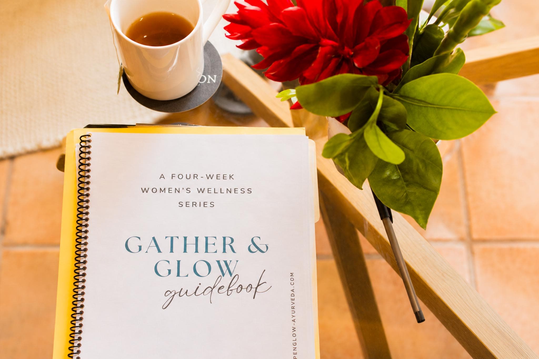 A wellness guidebook titled "Gather & Glow" is placed on a table next to a cup of tea and a vase of flowers.