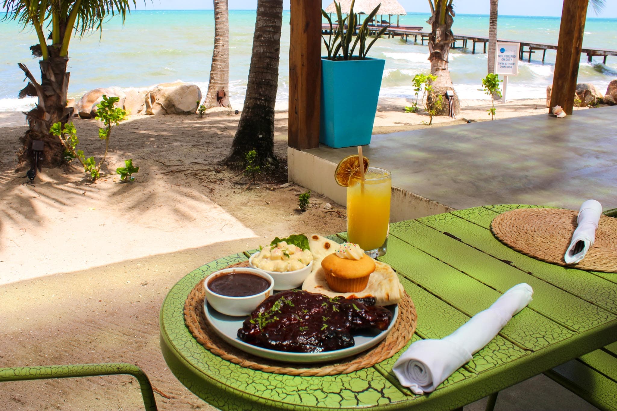A vibrant outdoor dining setting featuring a plate of barbecue chicken with sides and a refreshing drink against a scenic beach backdrop.
