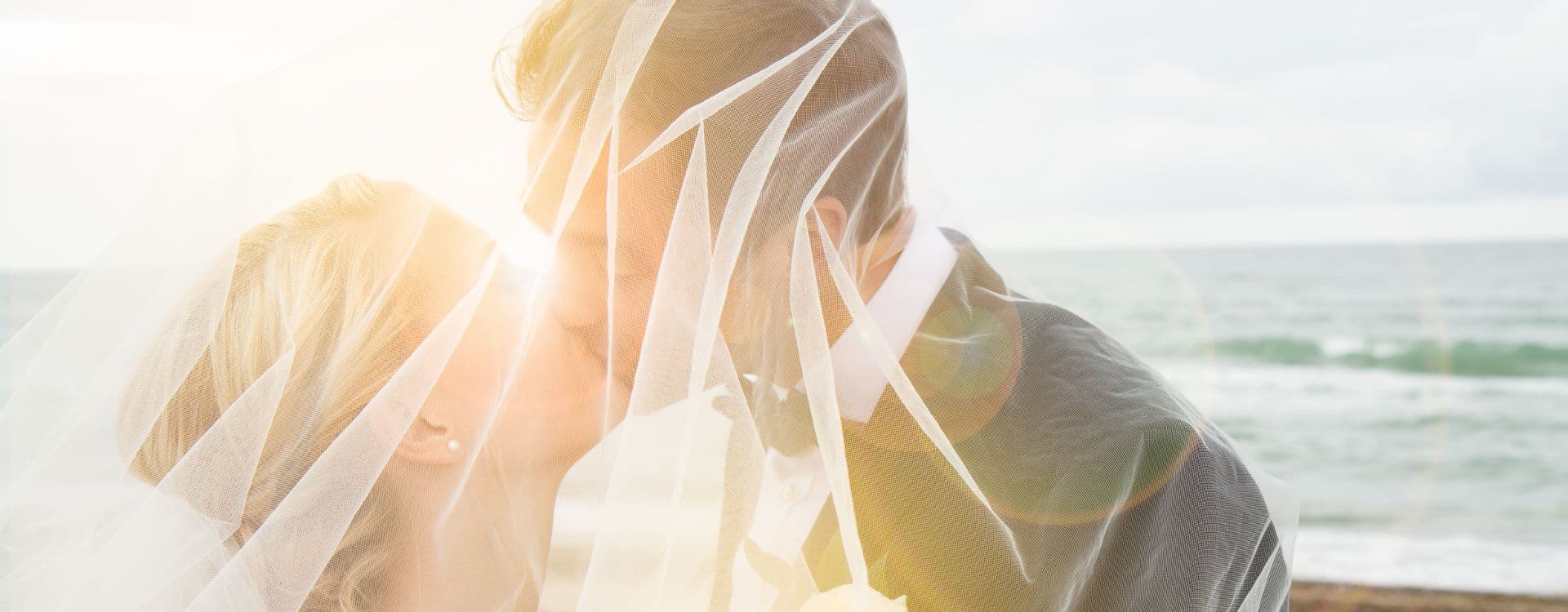 A couple kisses under a veil on a beach during sunset.