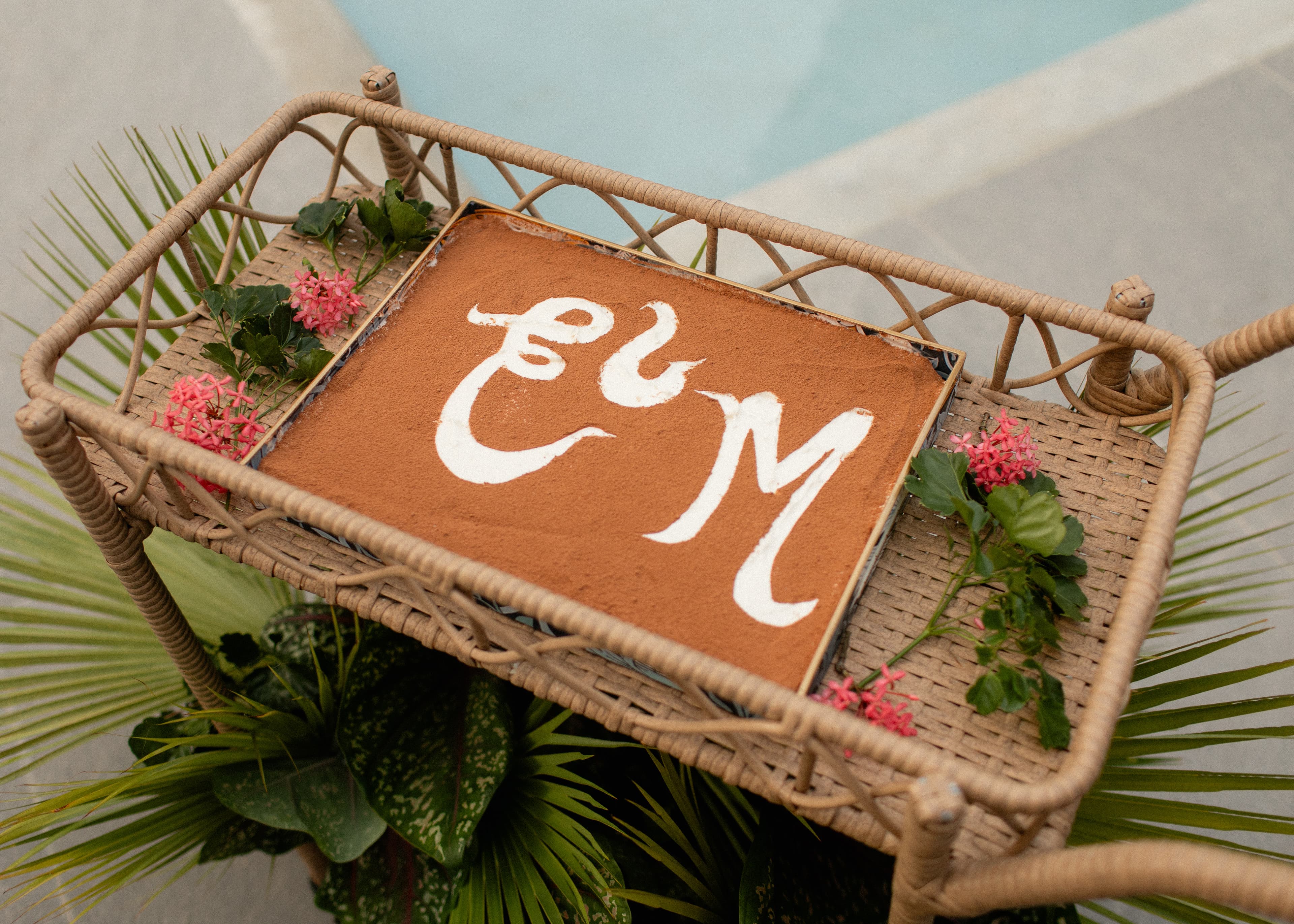A decorative basket with a brown surface featuring white artistic lettering and surrounded by greenery and small pink flowers.