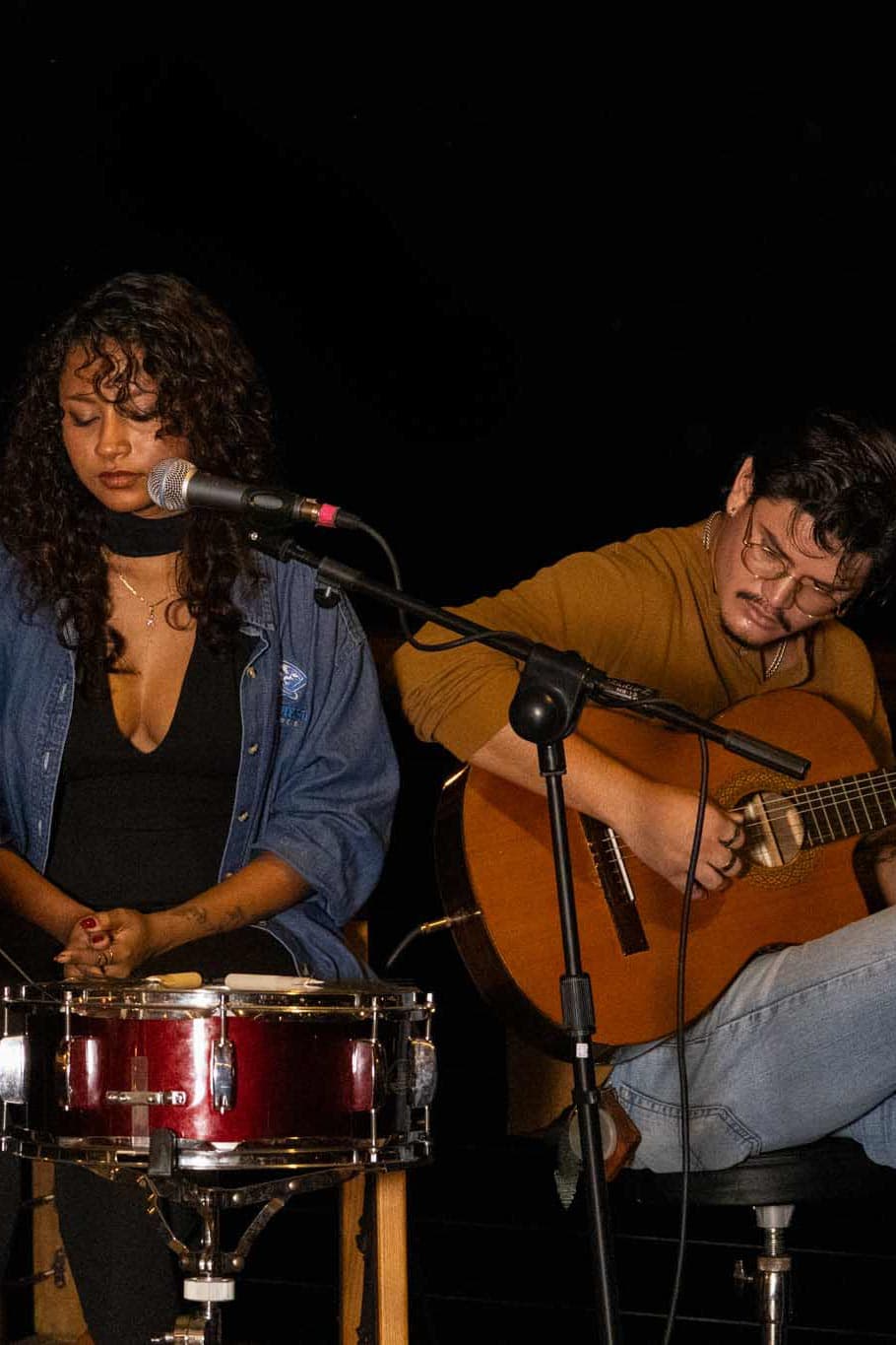 A musician plays guitar while another performs vocals and percussion on stage.