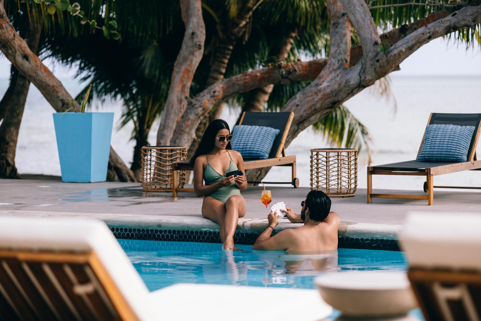 A woman in a swimsuit sits by a pool while a man enjoys a drink nearby.