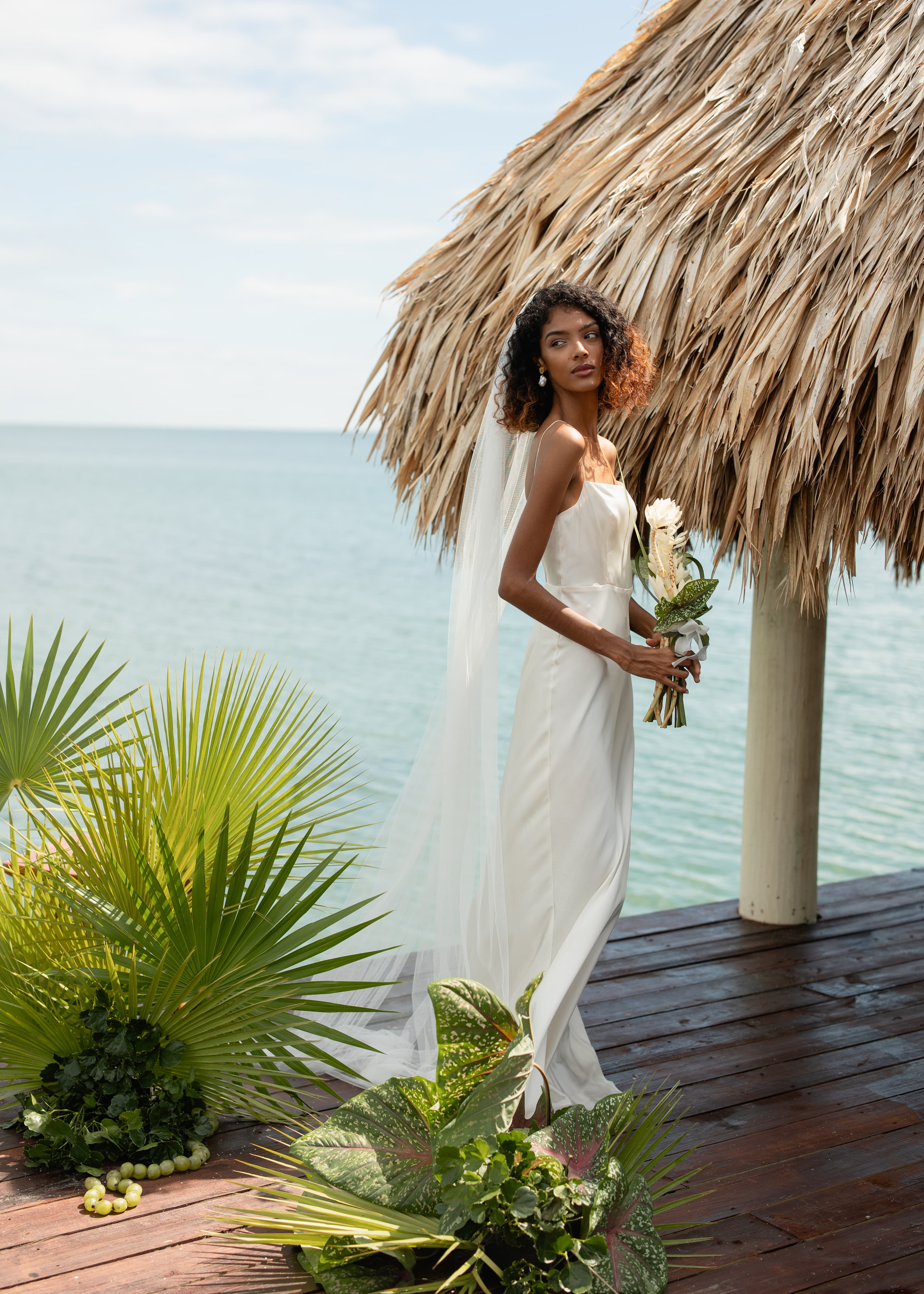 A bride in a flowing white dress holds a bouquet while standing on a wooden deck with tropical plants and a thatched structure by the sea.