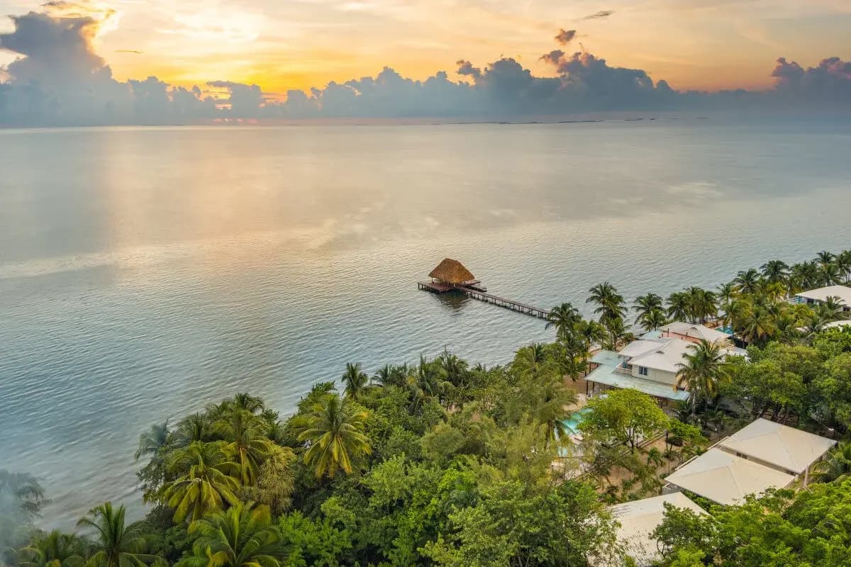 Sunset over calm waters with a thatched pier and lush tropical foliage.