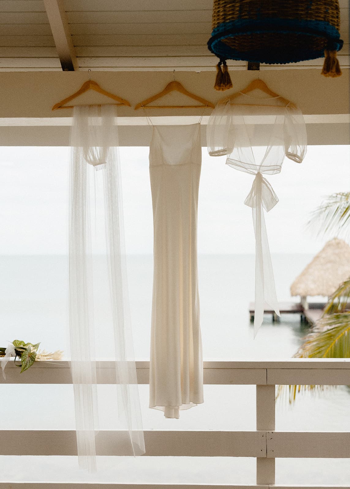 A white wedding dress and veil hang against a backdrop of serene water and a thatched-roof shelter.