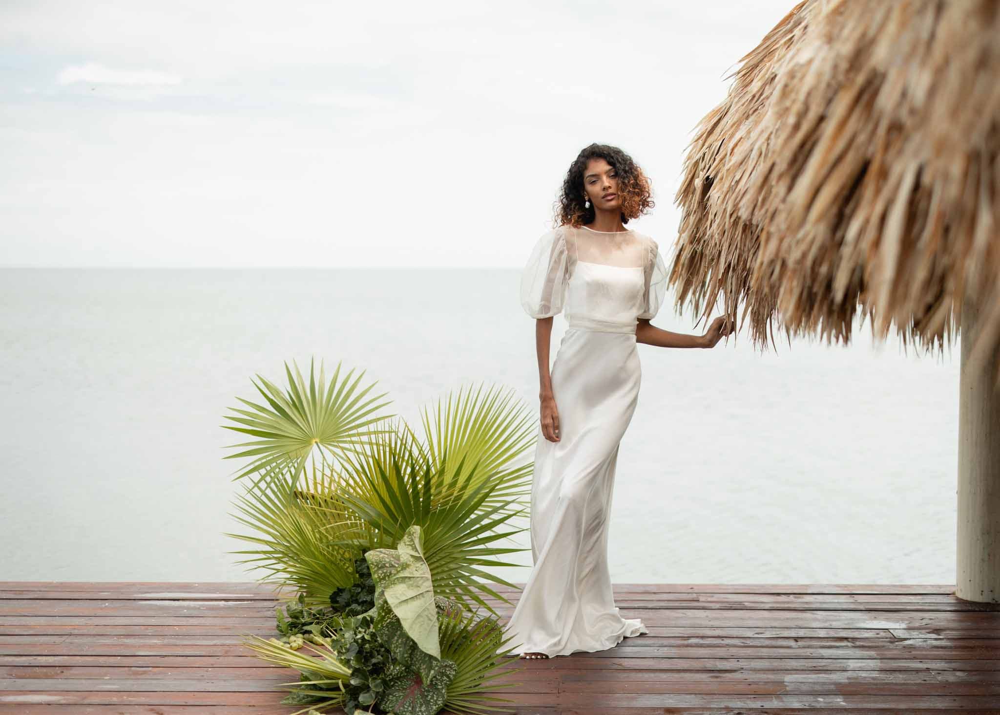 A woman in a flowing white dress stands on a wooden deck by the water, with tropical plants nearby and a thatched roof overhead.