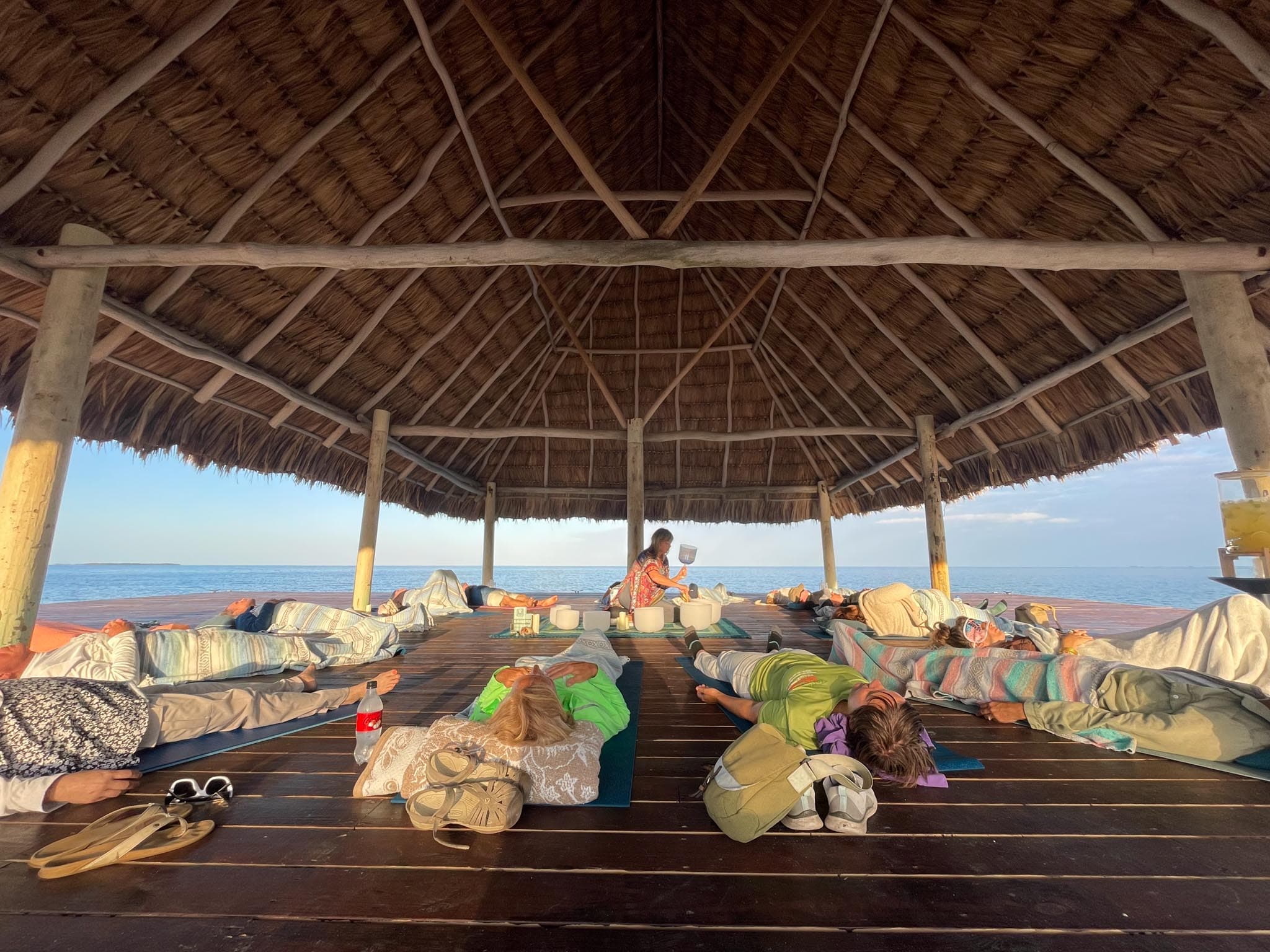A group of people is lying on mats in a thatched-roof structure by the sea.
