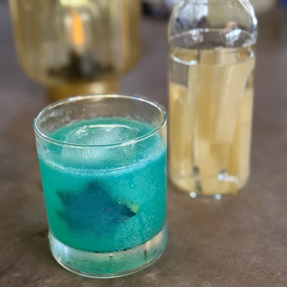 A vibrant blue cocktail sits in a glass next to a clear bottle of pale liquid.