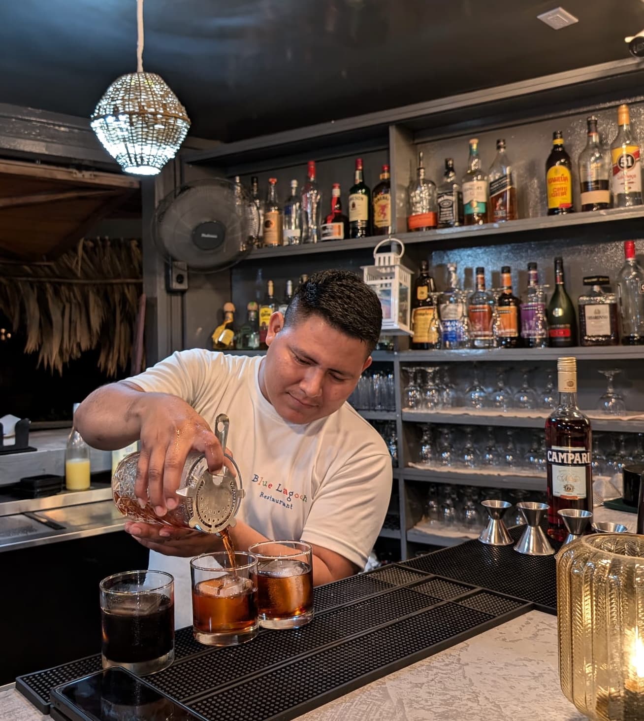 A bartender pours drinks into three glasses at a well-stocked bar.