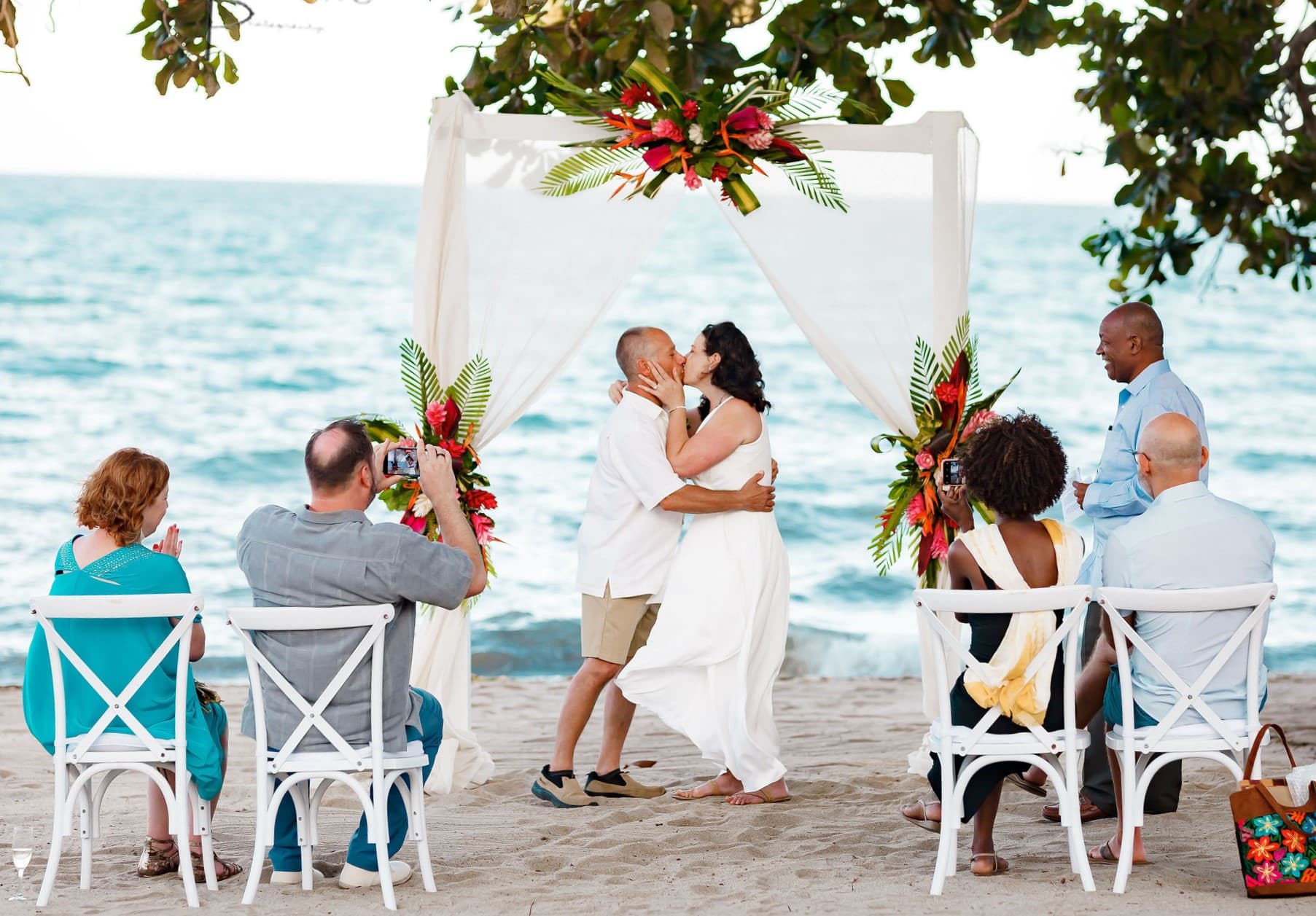 A couple kisses during their beachside wedding ceremony, surrounded by guests under a floral arch.