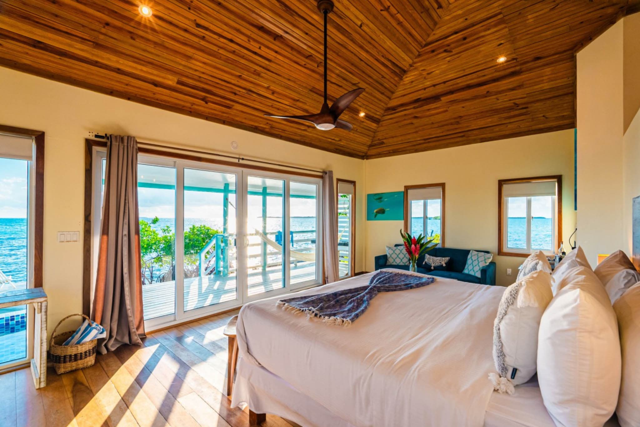 A bright, airy bedroom with wooden ceiling, large windows overlooking the ocean, and a neatly made bed.