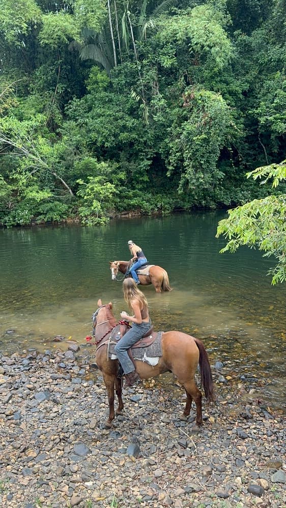 Two riders on horseback wade through a shallow river surrounded by dense greenery.