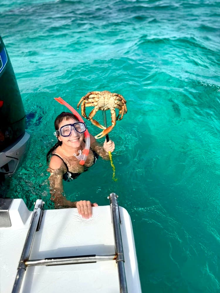 A smiling woman in snorkeling gear holds a crab while standing in clear turquoise water beside a boat.