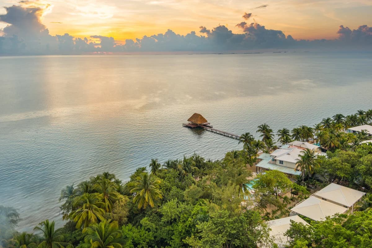 A tranquil sunset over calm waters with palm trees and a pier extending to a thatched gazebo.