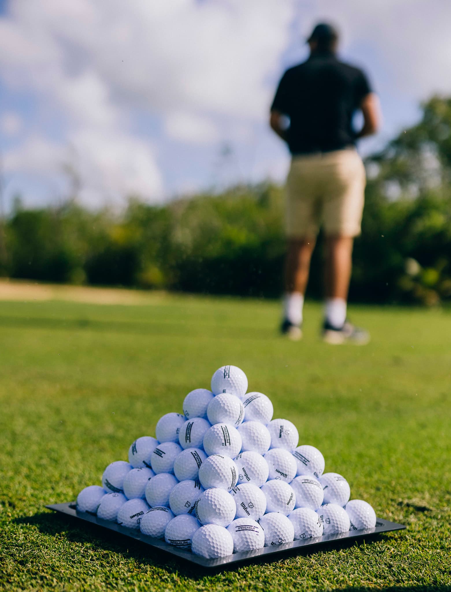 A pyramid of golf balls sits on the grass with a person in the background.
