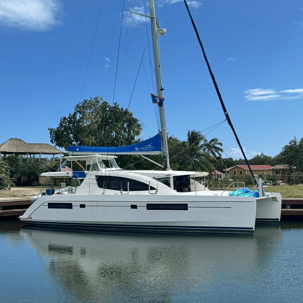 A white catamaran with a blue sail docked in a calm waterway under a clear blue sky.