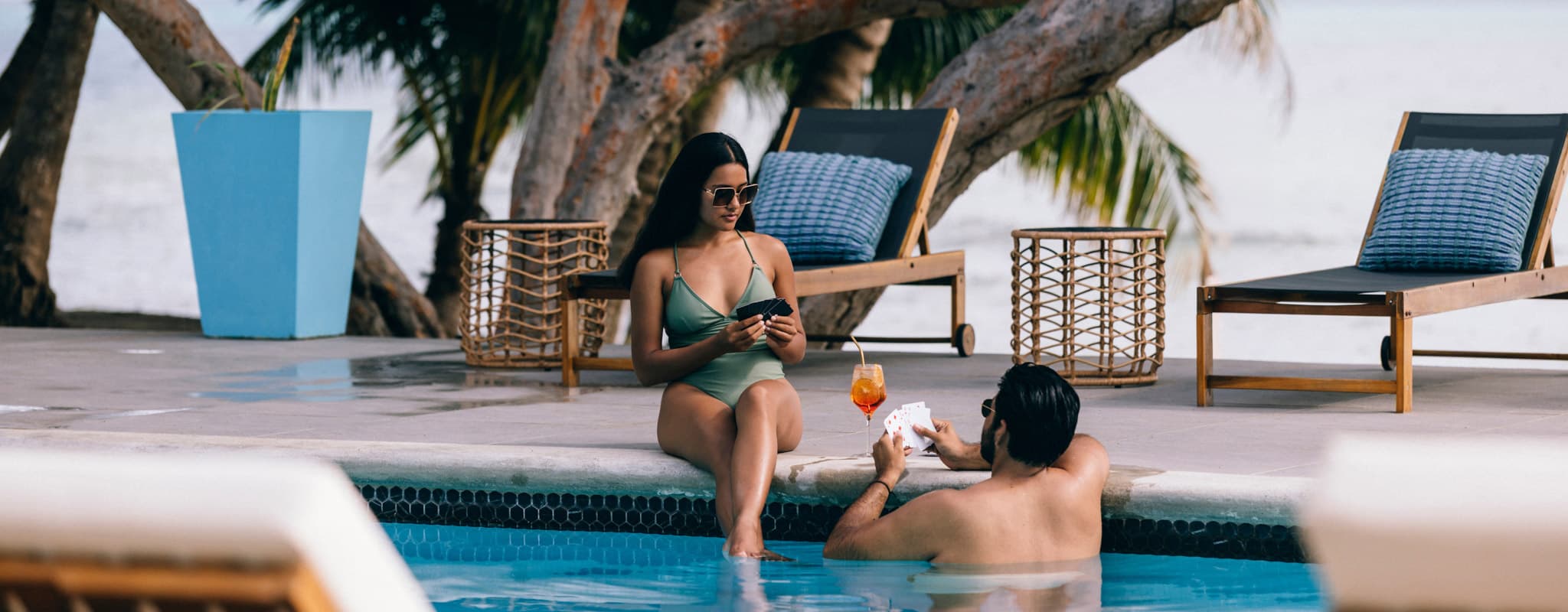 A woman in a green bikini sits by a pool while a man in the water enjoys a drink.