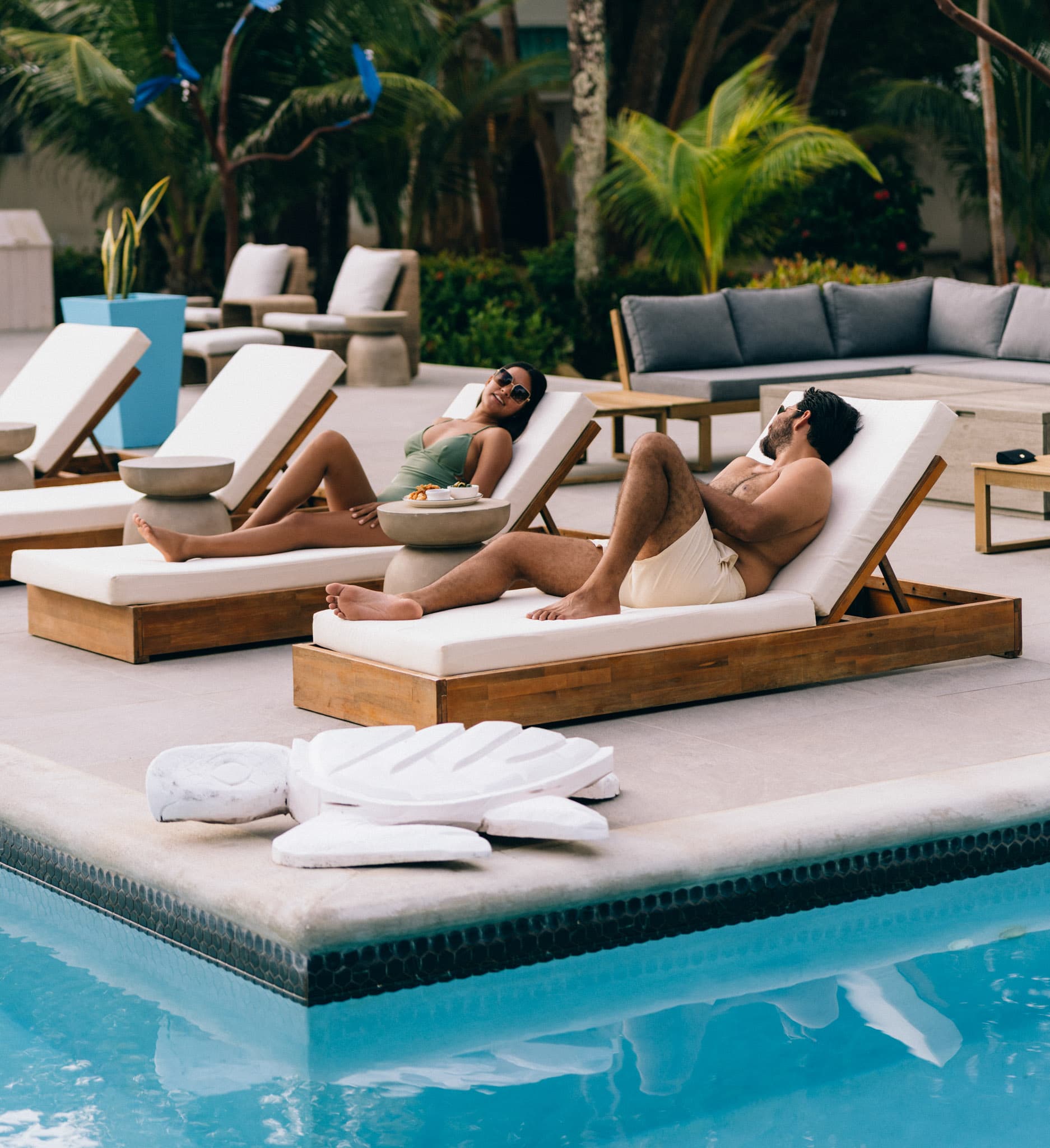 Two people relax on lounge chairs by a pool, with tropical greenery in the background.