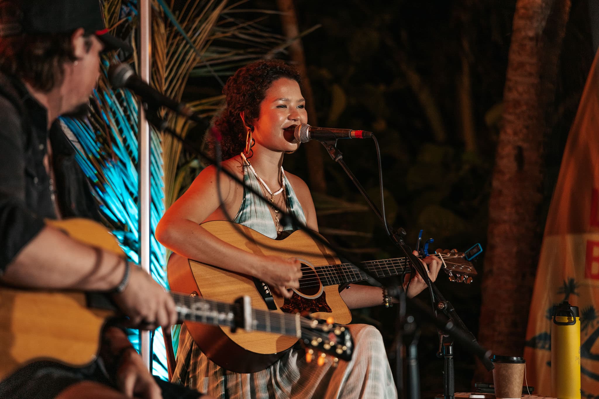 A woman with curly hair sings and plays an acoustic guitar while a man accompanies her on another guitar in a lively setting.