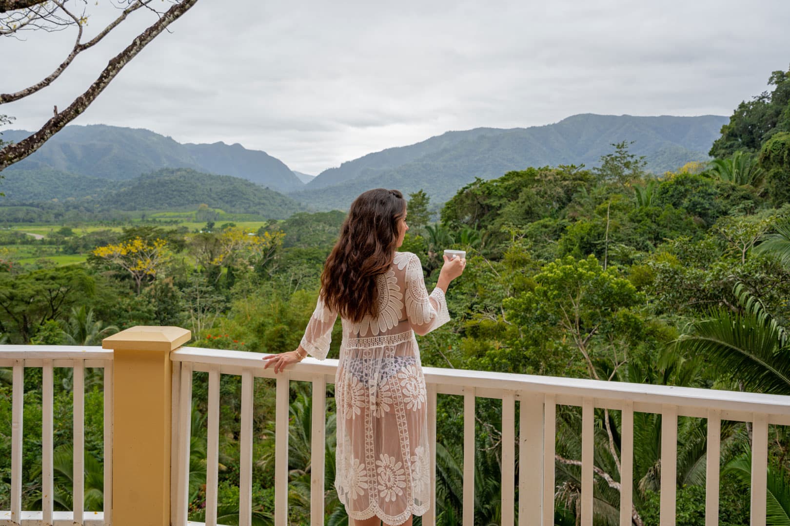 A woman in a sheer white dress stands on a balcony, gazing at a lush mountain landscape.