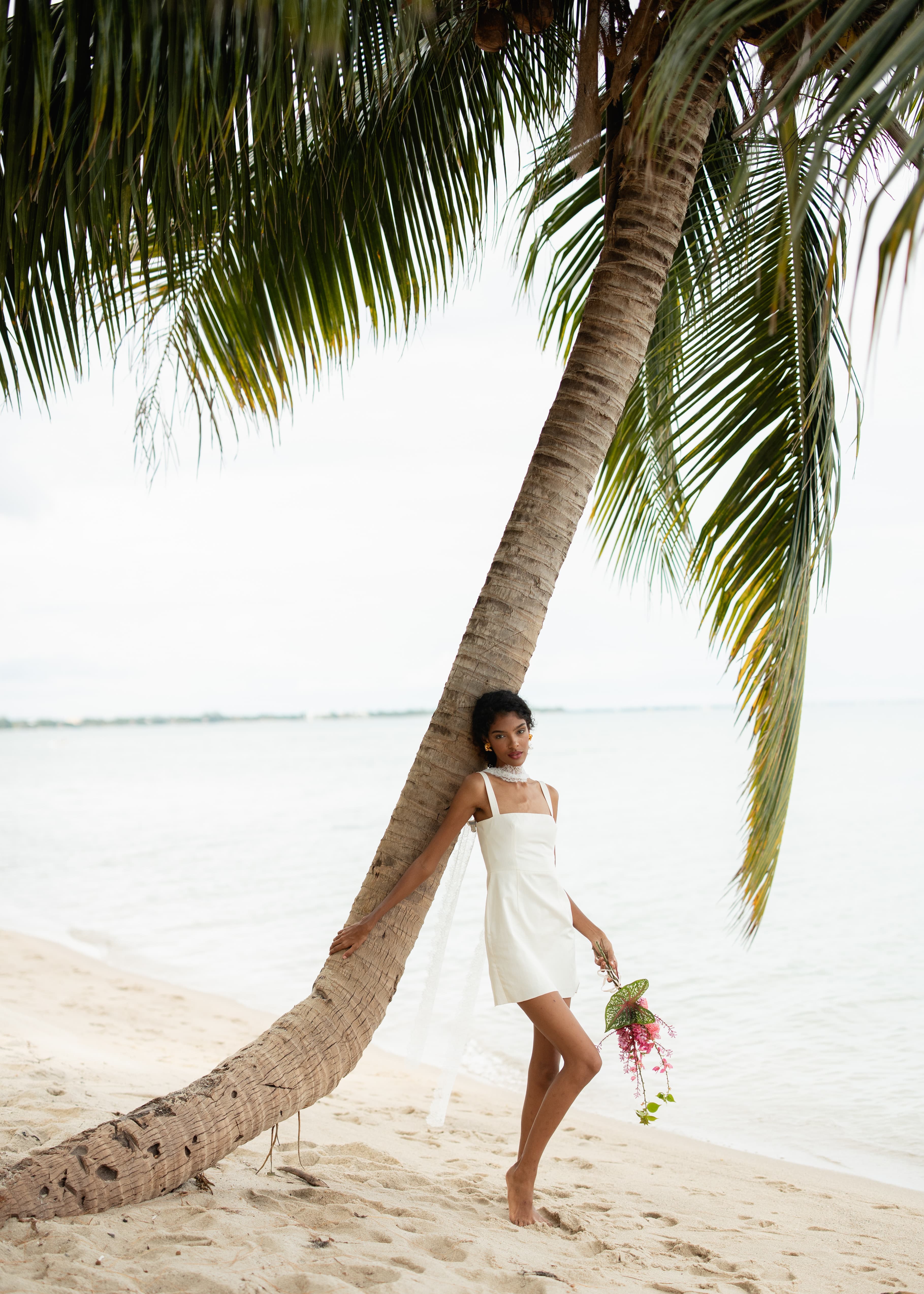 A woman in a white dress poses gracefully by a palm tree on the beach, holding a bouquet of flowers.