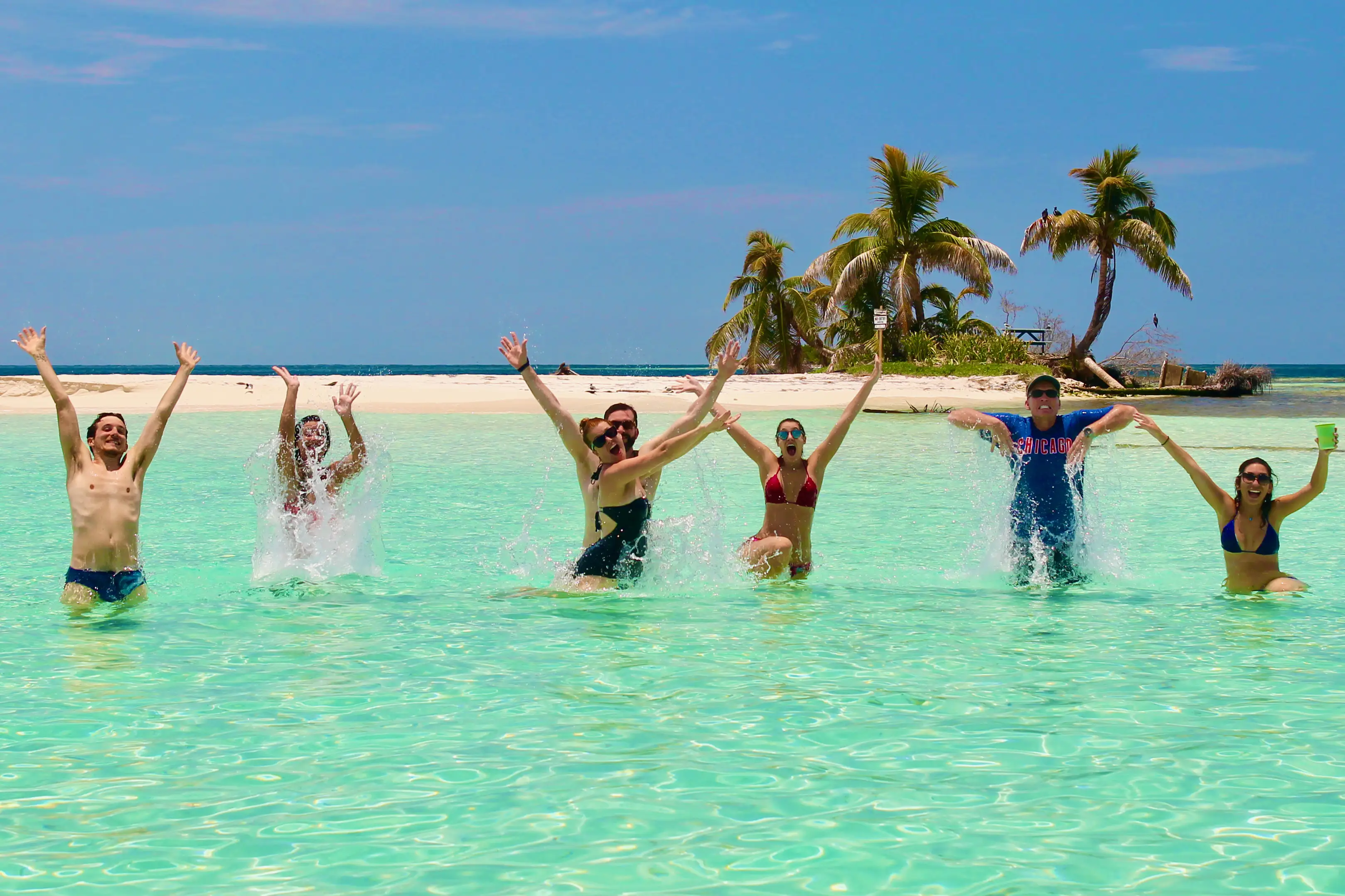 A group of six people joyfully splashes in clear turquoise water with a sandy beach and palm trees in the background.