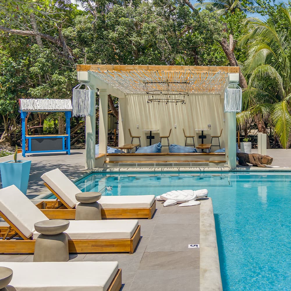 A serene poolside area featuring lounge chairs and a shaded seating space surrounded by lush greenery.