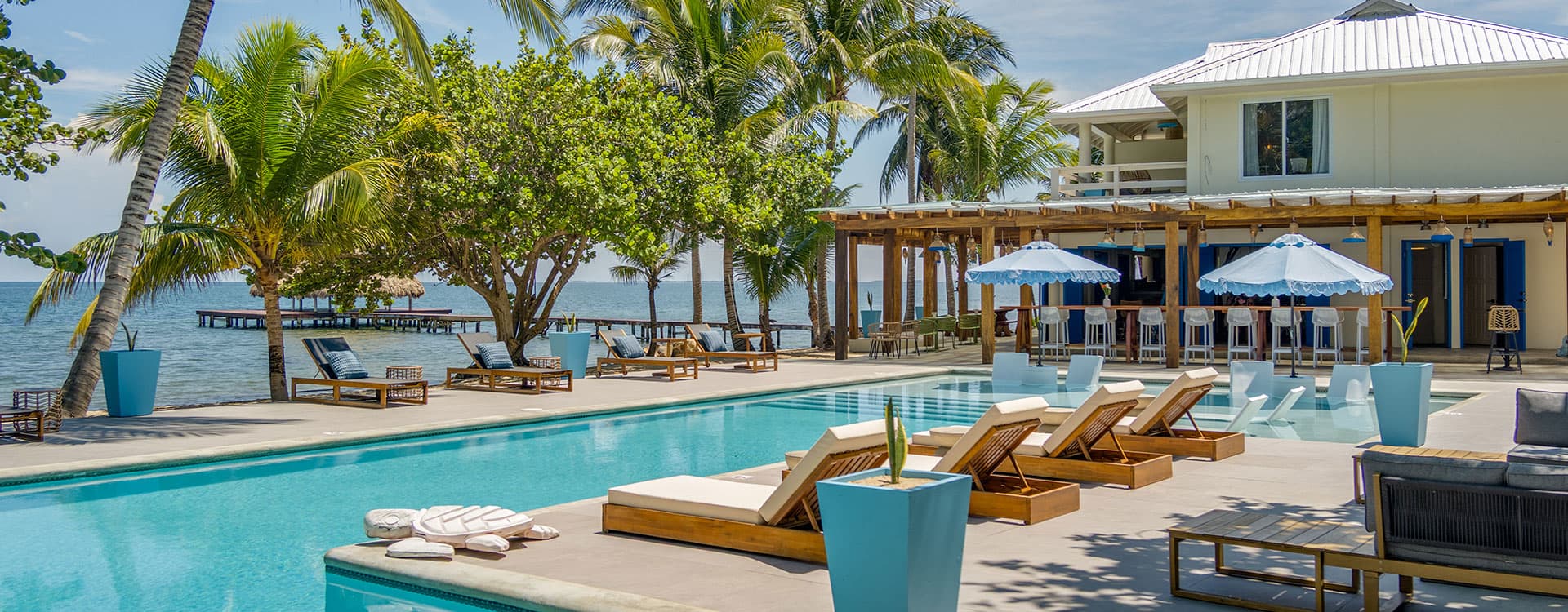 A sunny poolside view with lounge chairs, palm trees, and a waterfront house.