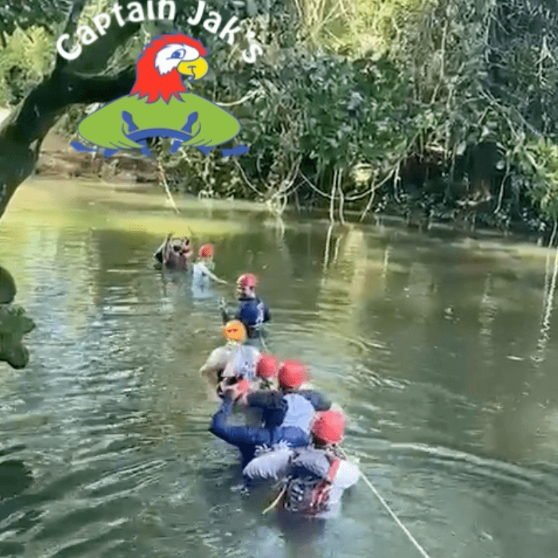 A group of people in helmets wade through a river while holding onto a rope, surrounded by lush greenery.