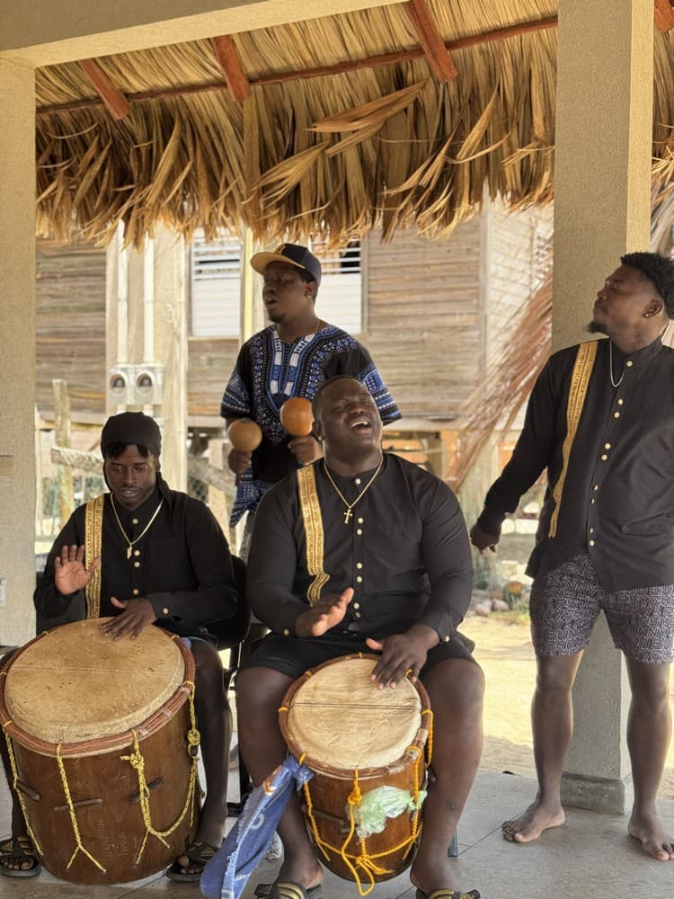 A group of four musicians performs with drums and maracas under a thatched roof.