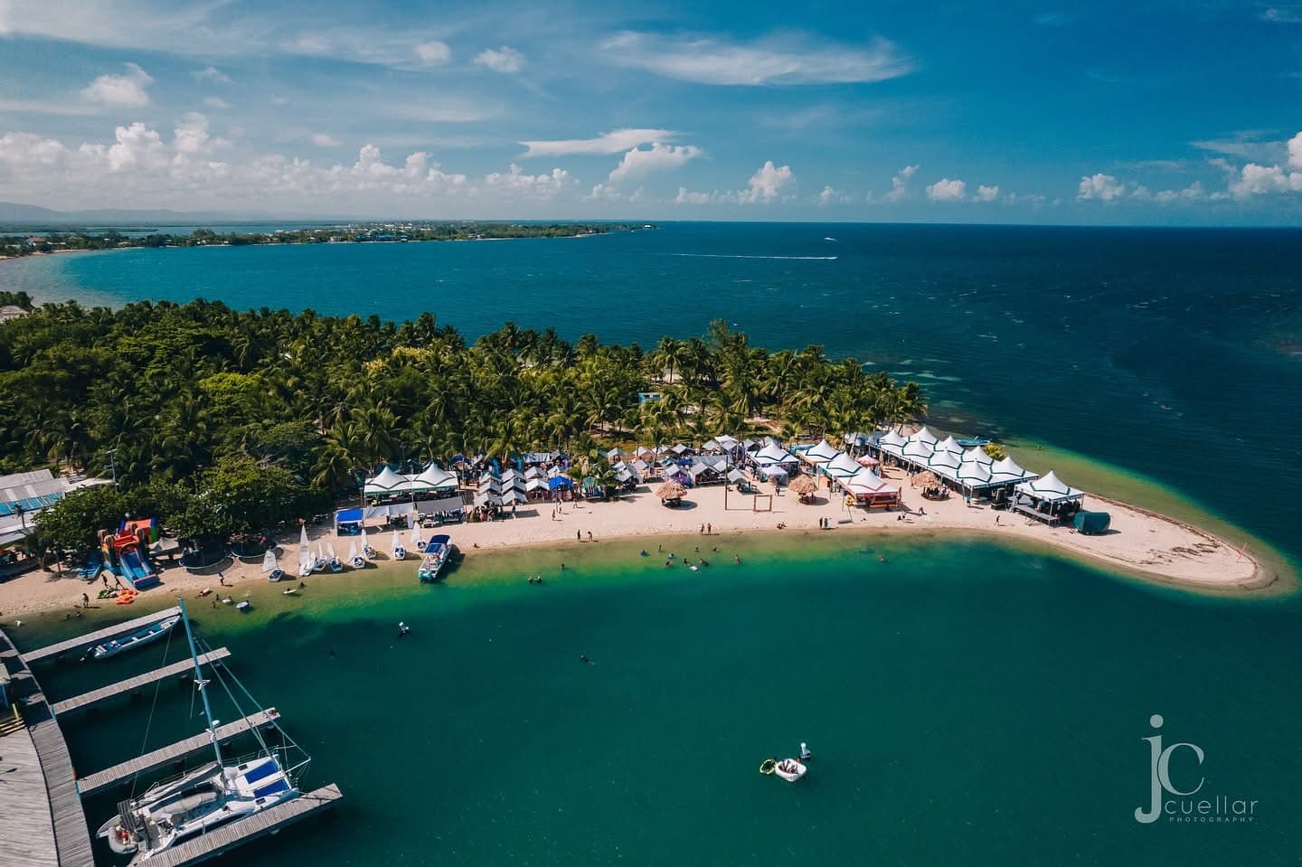 Aerial view of a lush, palm-fringed beach area with tents, people swimming, and clear blue waters.