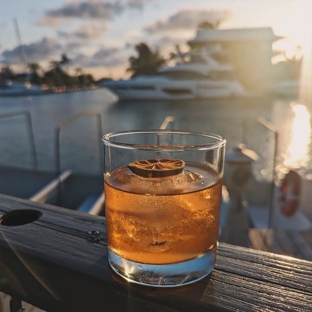 A glass of amber cocktail with a dried lime slice sits on a wooden railing, overlooking a marina at sunset.