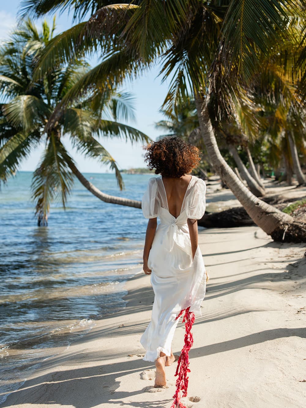 A woman in a white dress walks barefoot along a sandy beach lined with palm trees.