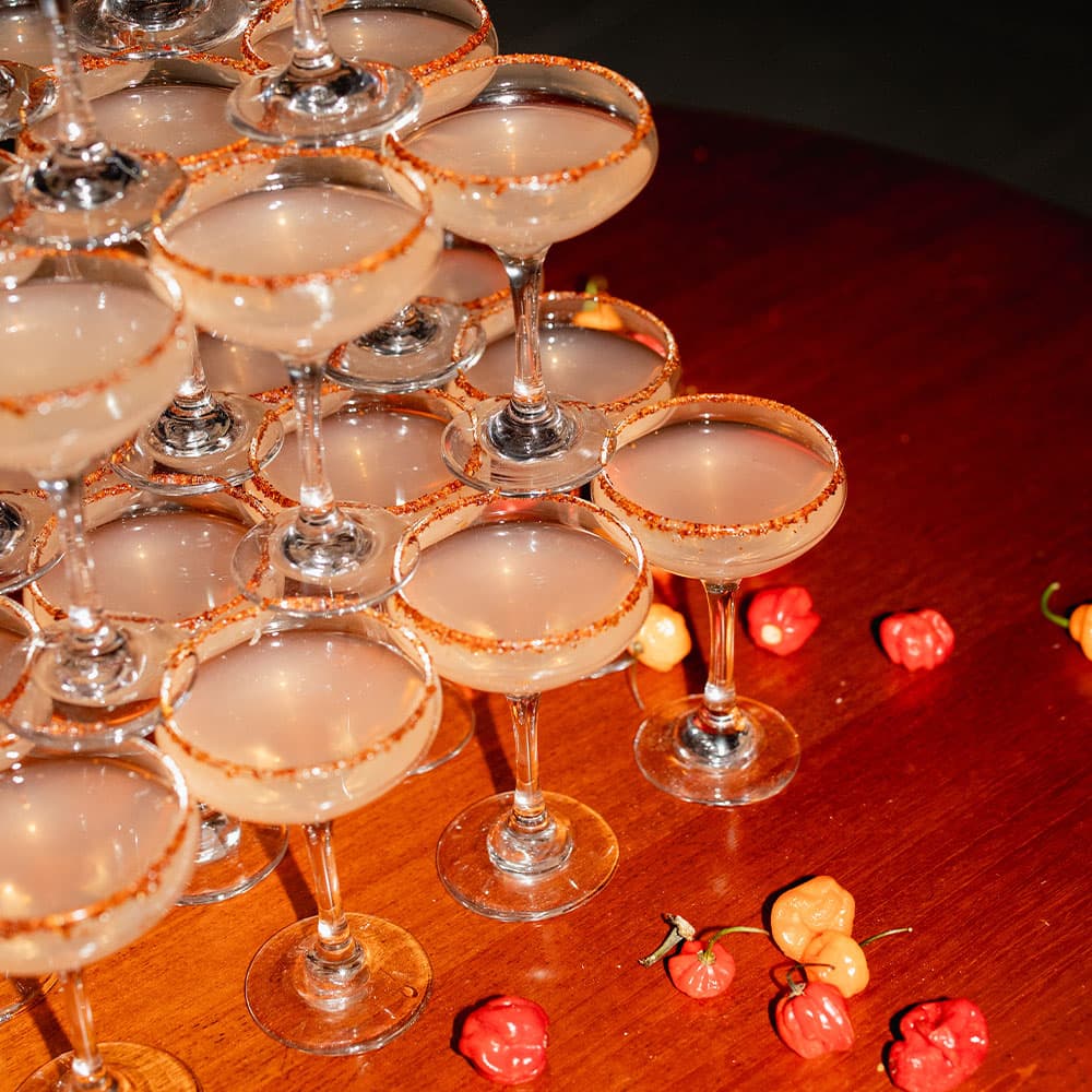 A pyramid of cocktails garnished with a rim of sugar sits on a wooden table, surrounded by small peppers.