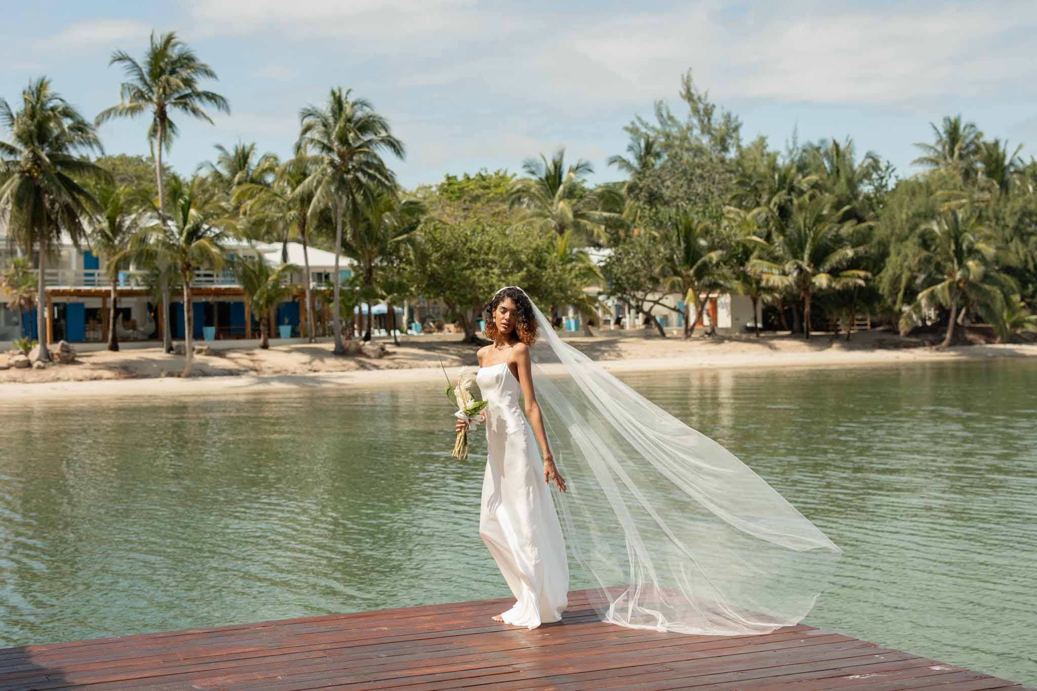 A bride in a flowing white gown and veil poses on a wooden dock beside a tranquil body of water, with palm trees in the background.