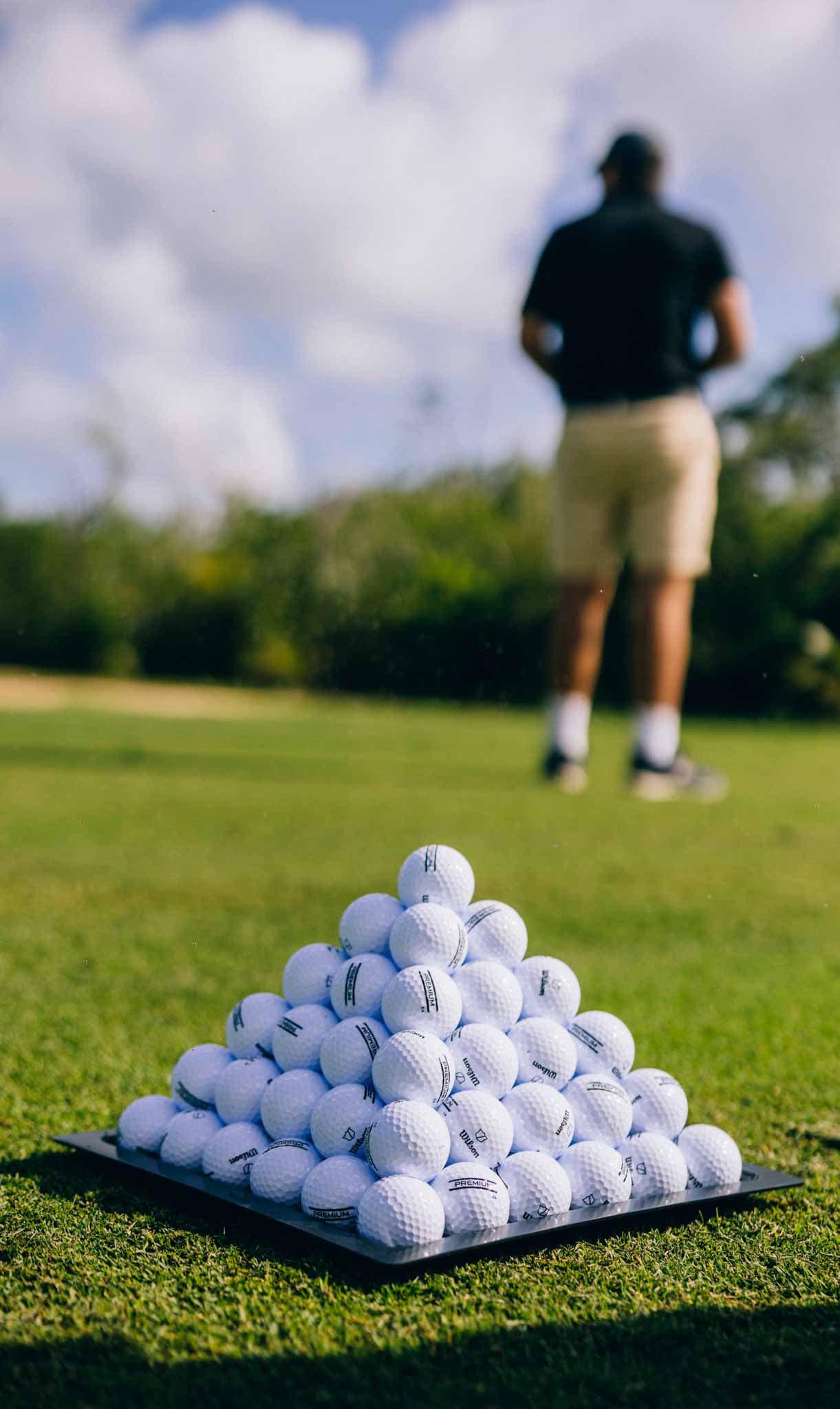 A pyramid of golf balls sits on the grass with a blurred figure in the background.