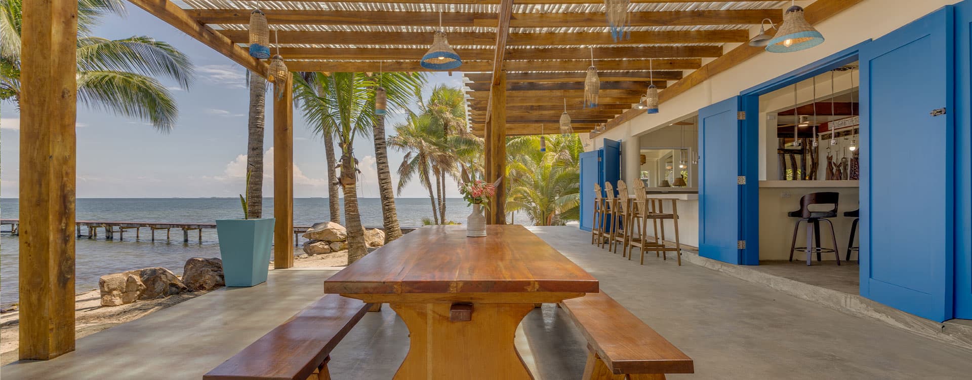 A rustic dining area with a wooden table under a thatched roof, overlooking the ocean and surrounded by palm trees.
