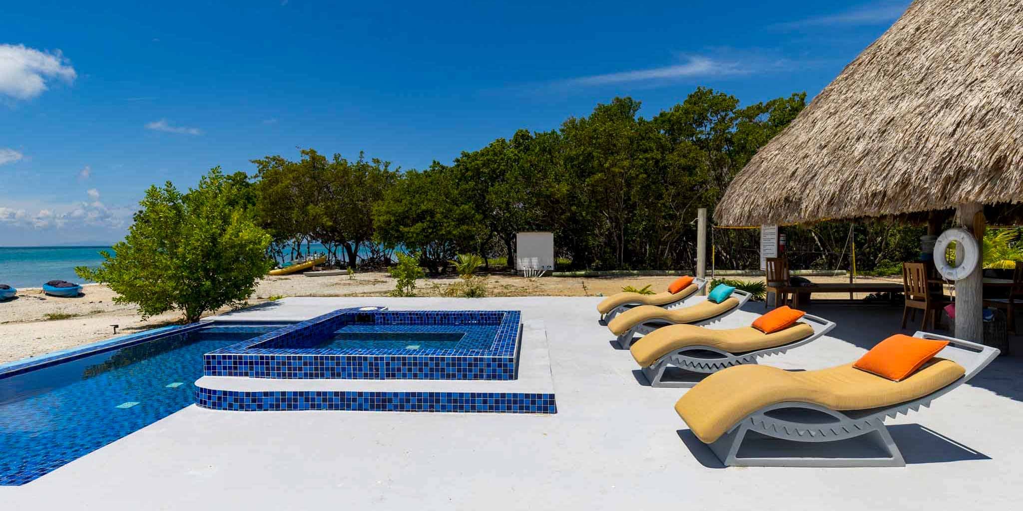 View of a beachside pool area with lounge chairs and a shaded cabana surrounded by greenery and ocean.