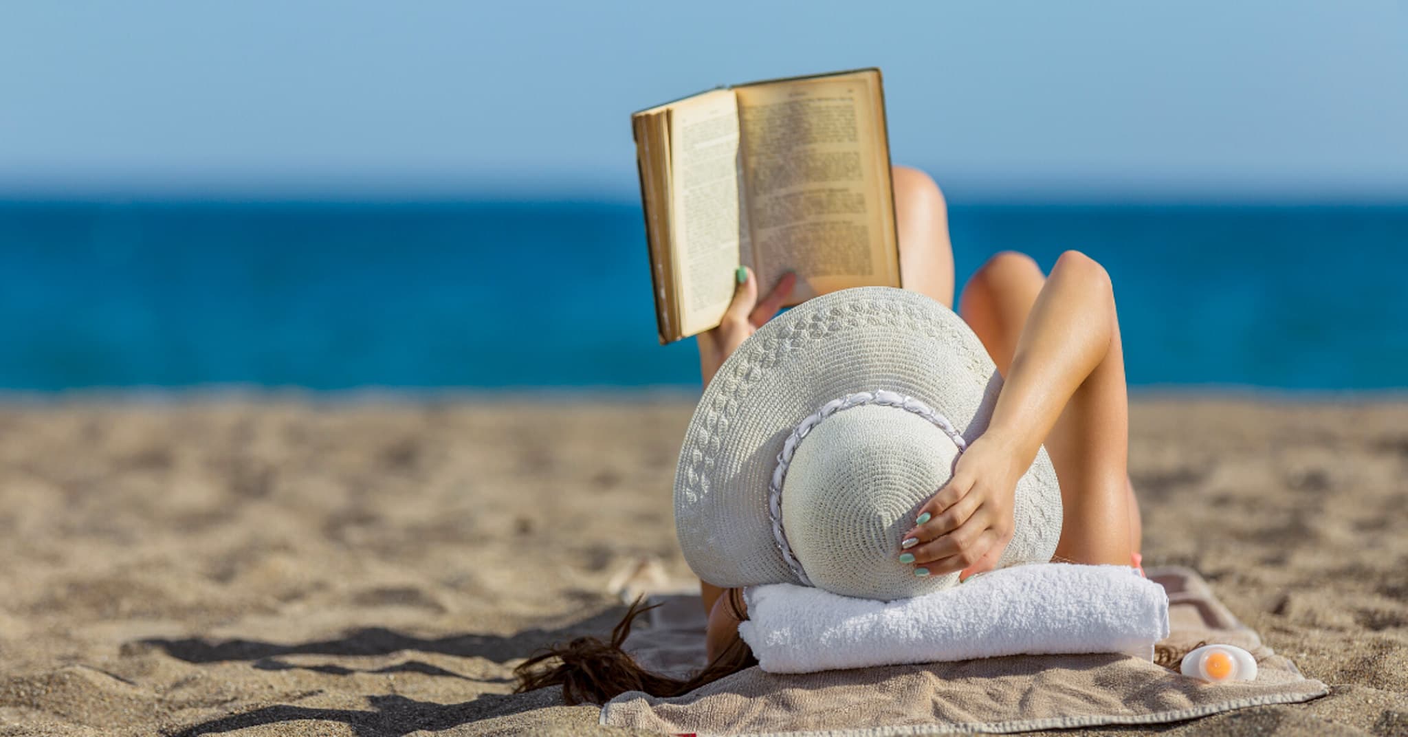A person relaxing on the beach, reading a book while wearing a wide-brimmed hat.
