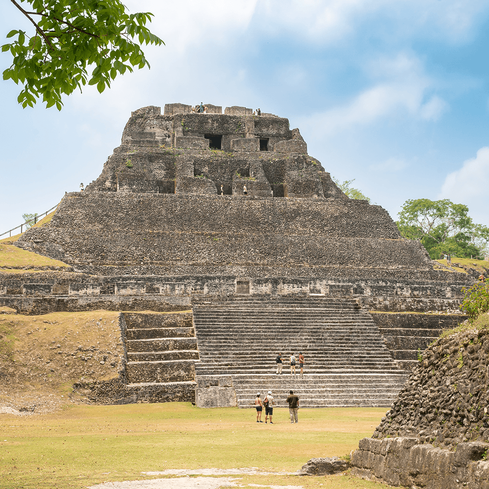 The image shows a large ancient pyramid with steps and people exploring around it under a blue sky.