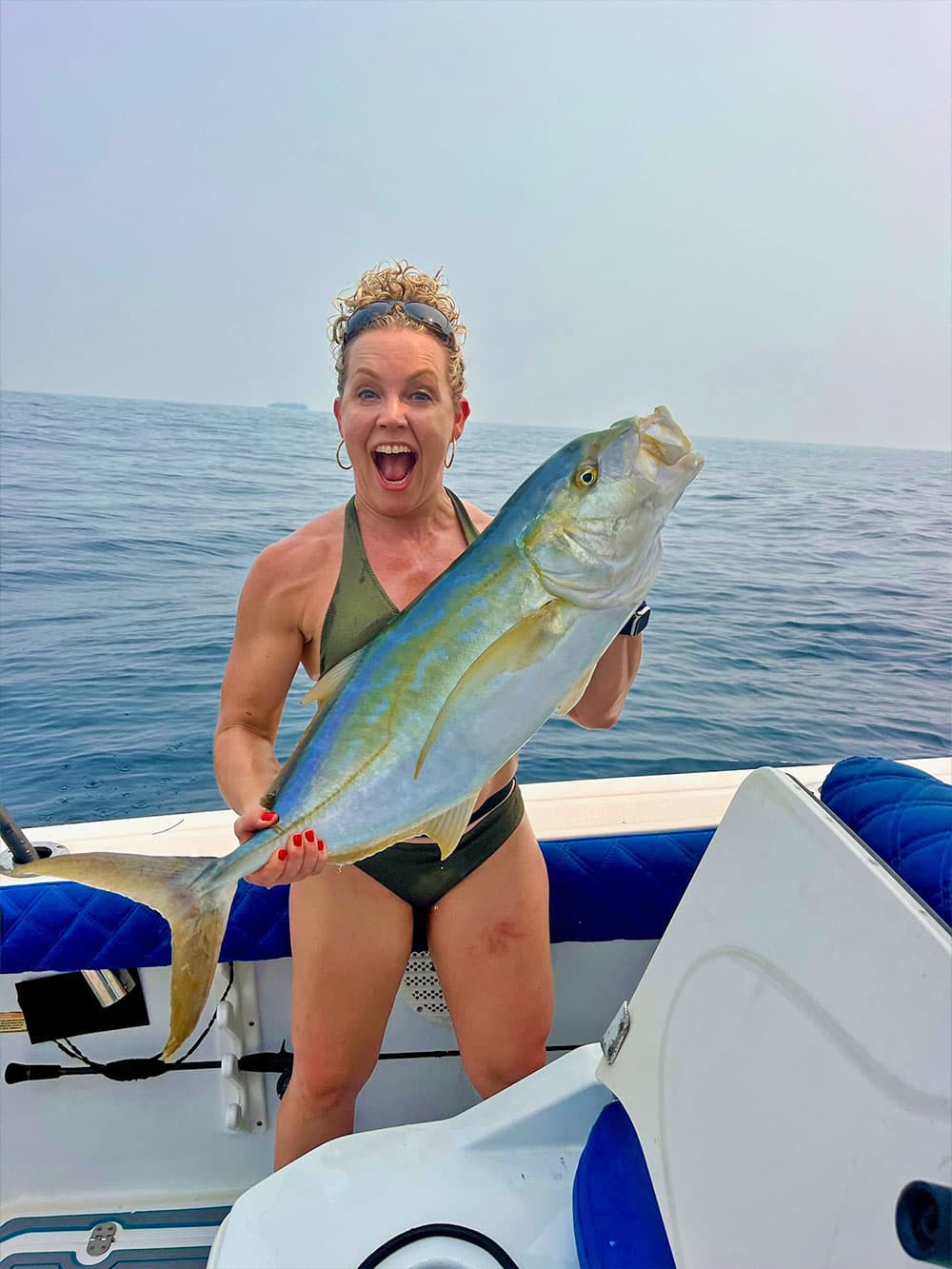 A woman joyfully holds a large fish while standing on a boat in the ocean.