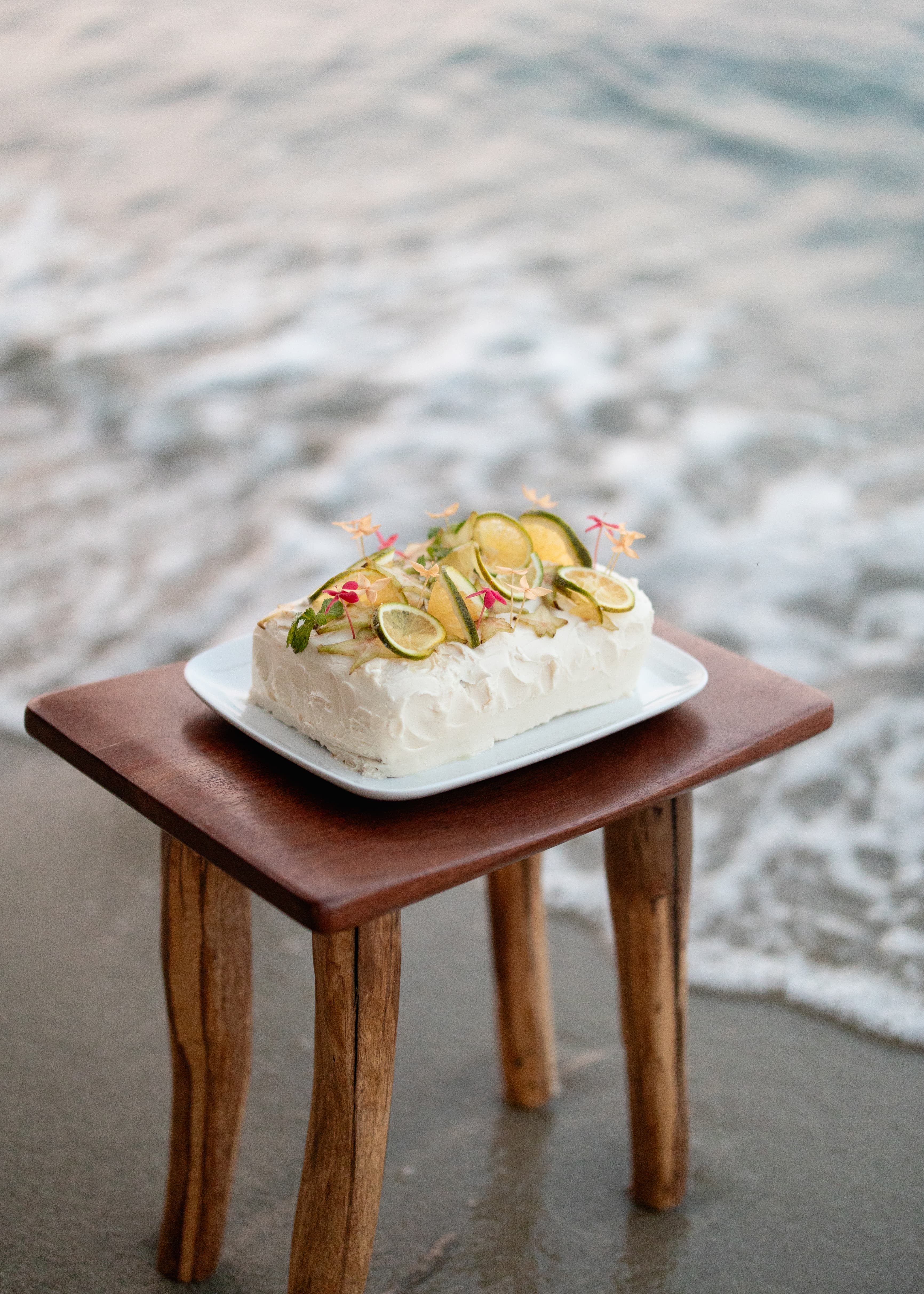 A decorated cake with lime slices and edible flowers sits on a wooden table by the shore.